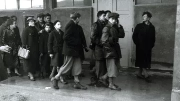 L'image montre un groupe de personnes, majoritairement des femmes et des enfants, marchant en direction d'un bâtiment. Ils portent des vêtements sombres et d'hiver. Leurs expressions semblent sérieuses et préoccupées. À l'entrée du bâtiment, une personne se tient à l'écart, observant le groupe avec une expression indéchiffrable. L'atmosphère de l'image est pesante et évoque un sentiment de tristesse ou d'incertitude. La scène semble avoir lieu dans un cadre urbain ou un lieu de rassemblement, avec des fenêtres en arrière-plan.