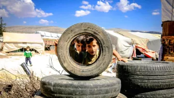 L'image montre deux enfants regardant à travers un pneu, dans un environnement qui semble être un camp de réfugiés. On peut voir des pneus empilés autour d'eux et des abris simples en arrière-plan. Le ciel est dégagé avec quelques nuages. Les enfants affichent une expression curieuse, et la scène dépeint un moment de jeu au milieu d'un cadre difficile.