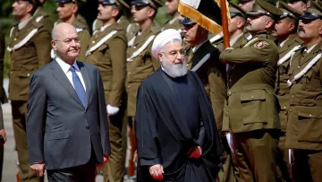 The image shows two men walking in a formal setting, likely during a ceremonial event. One man is dressed in a suit and tie, while the other is wearing a traditional black robe and a white turban. They are surrounded by a group of uniformed soldiers standing at attention, indicating a military or state occasion. The atmosphere appears formal and significant, suggesting a diplomatic meeting or official visit.