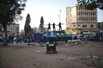 L'image montre une scène animée dans un endroit urbain. Au premier plan, on voit un espace dégagé avec un générateur. En arrière-plan, un certain nombre de personnes sont rassemblées, probablement pour assister à un événement. Sur le toit d'une fourgonnette bleue, trois personnes sont perchées, probablement en train de performer ou de s'exprimer de manière artistique. L'environnement semble être un mélange de vie urbaine et de rassemblement communautaire.