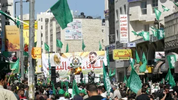 L'image montre une foule dense rassemblée dans une rue, manifestant avec des drapeaux verts. Au fond, il y a une grande banderole, probablement portant des messages politiques. Les gens semblent enthousiastes et engagés, et l'environnement est animé avec des bâtiments et des enseignes visibles autour. Les drapeaux verts peuvent indiquer l'association avec un mouvement ou un parti spécifique.