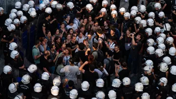 La imagen muestra una multitud de personas rodeadas por un grupo de policías. Los manifestantes parecen estar en una discusión o protesta, expresando sus opiniones entre ellos. Los policías, con cascos y uniformes, forman un cordón alrededor de la multitud, lo que sugiere una situación tensa. La escena parece tener lugar en un ambiente urbano, posiblemente durante una manifestación o conflicto social. La iluminación sugiere que puede ser al atardecer o al final del día.