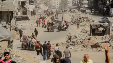 L'image montre une rue dévastée, probablement dans une zone de conflit. On peut voir des bâtiments en ruines de chaque côté de la voie, avec des débris éparpillés sur le sol. De nombreuses personnes, dont des familles et des individus, se déplacent sur la route. Certains utilisent des véhicules à trois roues, tandis que d'autres marchent à pied. L'atmosphère semble être celle d'une zone en reconstruction, avec beaucoup de poussière dans l'air et une ambiance de désolation.