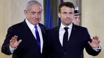 The image shows two men standing side by side, both dressed in formal attire. One is wearing a blue tie and the other has a dark tie, indicating a formal event. They appear to be waving, possibly to an audience or camera. The background suggests an official setting, likely an indoor venue with flags or formal decorations. Their expressions seem to convey a mix of seriousness and cordiality.