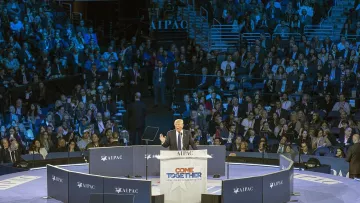 L'image montre un grand rassemblement dans une salle pleine de spectateurs. Au centre, une scène où un orateur, probablement un homme politique, s'exprime devant le public. Le logo "AIPAC" est visible sur des panneaux autour de la scène, indiquant que cet événement est lié à l'organisation pro-israélienne. Les participants semblent attentifs, et l'ambiance générale évoque un moment solennel et engagé. Les couleurs de l'éclairage sont principalement bleu et violet.