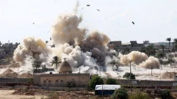 L'image montre une scène d'explosion avec de la fumée et des débris projetés dans les airs. En arrière-plan, on peut apercevoir des bâtiments, des palmiers et un terrain poussiéreux. L'explosion suggère un événement dramatique ou un conflit, et l'environnement semble rural ou semi-urbain. Des signes de destruction sont visibles, soulignant l'impact de l'explosion sur la zone environnante.