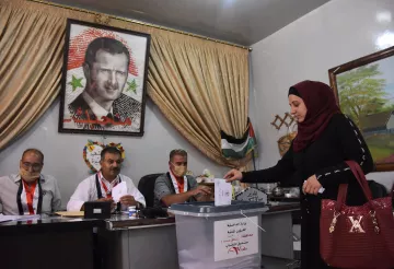 L'image montre une scène de vote, probablement lors d'élections en Syrie. Au premier plan, une femme en hijab dépose un bulletin dans une urne transparente. À l'arrière-plan, plusieurs hommes sont assis derrière une table, regardant le processus. Un grand portrait d'un homme, probablement un dirigeant, est accroché au mur, entouré de drapeaux et de décorations. Les participants portent des badges d'identification, et l'atmosphère semble sérieuse et formelle.