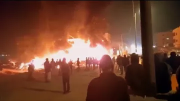L'immagine mostra una scena notturna in cui un edificio è avvolto dalle fiamme, creando una grande quantità di fumo e luce. Davanti all'edificio in fiamme, un gruppo di persone si raduna, osservando la situazione. Le figure sembrano essere in lontananza, mentre il fuoco illumina l'ambiente circostante. È possibile notare un'atmosfera di tensione e allerta.