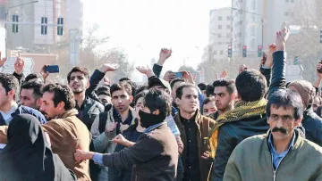 تظهر في الصورة مجموعة من الناس يتجمعون في مكان مفتوح، يبدو أنهم يشاركون في تظاهرة أو احتجاج. هناك مجموعة متنوعة من الأشخاص في الصورة، منهم رجال ونساء، وبعضهم يظهر بوجوه مكشوفة بينما البعض الآخر يلبس أغطية على وجهه. خلفهم يمكن رؤية لافتات وإشارات، مما يشير إلى أنهم يعبرون عن رأي أو موقف معين. الجو العام يوحي بالتحمس والجدل، حيث يبدو أن البعض يرفعون هواتفهم لتوثيق الحدث.