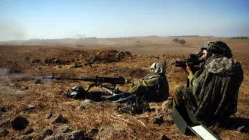 Sur l'image, on peut voir deux soldats en position de tir dans un paysage aride. Ils sont vêtus de tenue de camouflage et portent des casques. L'un des soldats est à genoux, prêt à tirer avec une mitrailleuse, tandis que l'autre est assis, observant à travers un dispositif de visée. Le terrain est composé de rochers et de terre sèche, et le ciel est dégagé, indiquant une journée ensoleillée. Cette scène évoque un environnement militaire austère et une préparation à l'action.