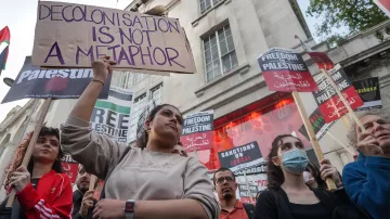 L'immagine mostra un gruppo di persone in una manifestazione, molte delle quali tengono in mano cartelli con frasi di protesta. Una giovane donna al centro tiene un cartello che dice "La decolonizzazione non è una metafora". Sullo sfondo, ci sono altri cartelli con scritte come "Libertà per la Palestina" e "Sanzioni a Israele". I partecipanti sembrano esprimere il loro sostegno per la causa palestinese, mentre l'atmosfera è di attivismo e impegno sociale.