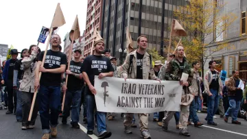 L'image montre un groupe de manifestants marchant dans une rue, tenant des pancartes. Au premier plan, plusieurs personnes portent des t-shirts noirs avec des slogans, et l'un d'eux tient une grande banderole qui dit "Iraq Veterans Against The War". Il semble s'agir d'une manifestation contre la guerre en Irak, avec des participants d'âges et d'apparences variés. L'ambiance semble sérieuse et engagée. Des bâtiments urbains et des arbres aux feuilles jaunes sont visibles en arrière-plan, suggérant que la scène se déroule en automne.