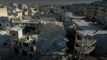 L'image montre un paysage urbain dévasté, probablement dans une zone de conflit. On y voit des bâtiments en ruines, certains complètement effondrés, d'autres partiellement intacts. Le sol est jonché de débris, témoignant des destructions causées par des bombardements ou des combats. L'environnement paraît déserté, avec peu ou pas de signes de vie, illustrant la tragédie et les conséquences dramatiques de la guerre sur les infrastructures urbaines.