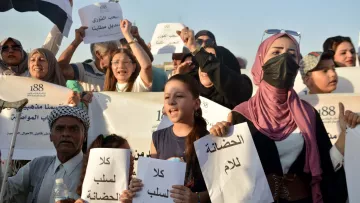 L'image montre un groupe de personnes lors d'une manifestation. Des manifestants, y compris des enfants, tiennent des pancartes avec des inscriptions écrites en arabe. Ils expriment des revendications, visiblement engagés dans une lutte pour une cause qui leur tient à cœur. Certaines personnes portent des masques, et l'ambiance semble déterminée et pleine d'énergie. Le paysage semble ensoleillé, suggérant un rassemblement en plein air.