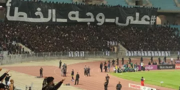 Stade plein de supporters en noir avec une grande banderole en arabe. Énergie et passion.