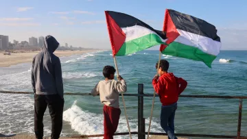 L'image montre trois enfants debout sur un bord de mer, regardant vers l'horizon. Deux des enfants tiennent des drapeaux avec les couleurs noir, rouge, vert et blanc, emblématiques de la Palestine. La mer est agitée, avec des vagues qui s'écrasent contre le rivage, et en arrière-plan, on peut apercevoir des bâtiments sur la plage. Les enfants semblent contemplatifs, profitant de l'instant. Le ciel est partiellement nuageux, créant une atmosphère tranquille.