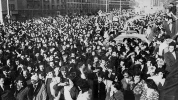 L'image montre une grande foule rassemblée dans une rue. Les personnes semblent se tenir très proches les unes des autres, créant une masse compacte. On peut apercevoir des bâtiments en arrière-plan, ainsi que des véhicules, ce qui indique que cette scène se déroule dans un milieu urbain. L'atmosphère semble intense et collective, suggérant un rassemblement pour une cause ou un événement significatif. Les vêtements des personnes, sombres et variés, donnent une impression de l'époque.