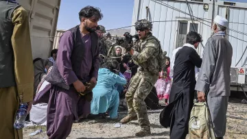 L'image montre une scène d'une situation tendue, probablement dans un contexte de conflit. On peut y voir un soldat en uniforme, armé et préparé, qui semble interagir avec d'autres personnes. Plusieurs civils sont présents, certains assis par terre, d'autres en mouvement, ce qui indique une atmosphère d'urgence. L'environnement est dénudé avec des bâtiments en arrière-plan ressemblant à des conteneurs. Les vêtements traditionnels des civils suggèrent qu'ils pourraient venir d'une région du Moyen-Orient ou d'Asie centrale. L'ensemble de la scène reflète une situation complexe et potentiellement dramatique.