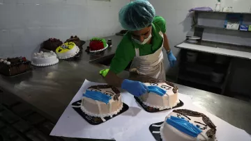 L'image montre un pâtissier en train de décorer des gâteaux dans une cuisine professionnelle. Il porte un bonnet de protection et des gants en latex. Sur la table, plusieurs gâteaux sont disposés, chacun ayant une décoration distincte, notamment des motifs qui ressemblent à des visages portant des masques. L'ambiance est soignée et les gâteaux sont présentés sur des plateaux noirs.