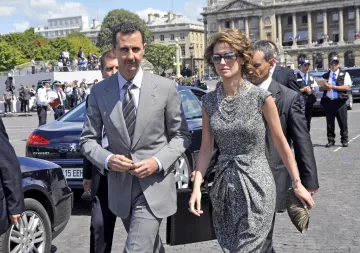 The image shows a man and a woman walking together in an outdoor setting. The man is dressed in a grey suit and tie, while the woman is wearing a stylish, patterned dress, sunglasses, and holds a handbag. They appear to be in a public area, possibly during a formal event, with a group of people in the background, including security personnel. The architecture behind them suggests a significant building or landmark.