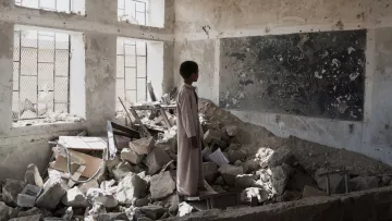 L'image montre un enfant se tenant dans une salle de classe en ruines. Le sol est jonché de débris, tandis que des morceaux de murs effondrés et des meubles renversés témoignent de la destruction. Un tableau noir, partiellement visible, porte des inscriptions, bien que l'environnement soit dégradé. La lumière pénètre à travers les fenêtres, créant un contraste avec l'atmosphère sombre et désolée de la pièce. Cette scène évoque une profonde sensation de perte et de désespoir.