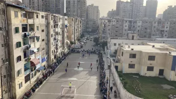 L'image montre un terrain de football encadré par des immeubles d'apartements dans une zone urbaine. Plusieurs personnes sont rassemblées sur des bancs le long du terrain, probablement en train de regarder un match. On peut voir des joueurs en action sur le terrain. L'établissement est entouré de bâtiments de plusieurs étages, témoignant d'une ambiance animée et communautaire. L'éclairage du soir ajoute une atmosphère chaleureuse à la scène.