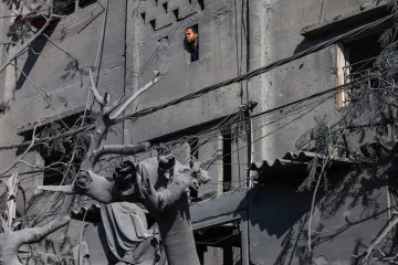 L'image montre un bâtiment en ruines avec des murs de béton gris, où l'on aperçoit une fenêtre. À travers cette fenêtre, une personne regarde vers l'extérieur. Au premier plan, on peut voir des débris et une statue partiellement détruite, entourée de fils électriques et de restes de végétation, ce qui donne une impression de désolation. L'environnement semble touché par un événement tragique, avec une atmosphère sombre et sombre.