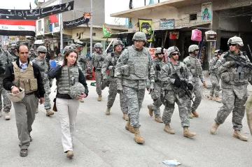 L'image montre un groupe de soldats en uniforme militaire marchant dans une rue. Ils semblent être dans une zone urbaine, possiblement dans un pays en conflit. Au premier plan, on voit une femme en vêtements civils, portant un gilet pare-balles, marchant à côté d'un homme également en tenue civile. Le décor autour d'eux comprend des bâtiments, des panneaux et d'autres signes de vie urbaine. L'atmosphère semble sérieuse, reflétant un environnement potentiellement tendu.