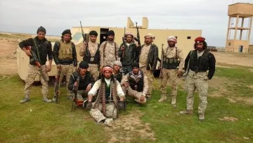 L'image montre un groupe de personnes en tenue militaire, armées et posant devant un véhicule. Ils semblent être sur un terrain en extérieur, peut-être dans un environnement désertique ou rural. Les hommes affichent diverses expressions et postures, certains sont debout tandis que d'autres sont assis. Leurs vêtements et accessoires suggèrent qu'ils font partie d'une unité militaire ou paramilitaire. En arrière-plan, on aperçoit un bâtiment et une tour d'eau.