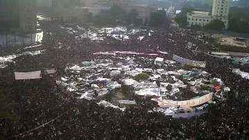 Una grande folla si raduna in una piazza, con tende e striscioni.