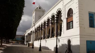 L'image montre un bâtiment architectural imposant, probablement un hôtel de ville ou un bâtiment administratif, avec une façade blanche. Ce bâtiment présente des arches décoratives et des fenêtres ornées. On peut apercevoir une grande horloge sur la tour, ainsi qu'un drapeau turc flottant au sommet. Le ciel est nuageux, ajoutant une ambiance dramatique à la scène. Des lampadaires sont alignés le long de la rue pavée qui borde le bâtiment, et un vélo est garé sur le côté. Des arbres bien taillés sont également visibles, contribuant à l'esthétique générale de l'endroit.