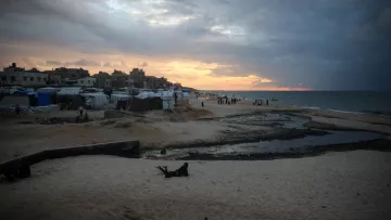 L'immagine mostra una spiaggia al tramonto, con il cielo che presenta nuvole scure e toni arancioni. Sulla spiaggia si trovano diverse tende e strutture, probabilmente utilizzate come rifugi temporanei. L'acqua del mare appare calma, e ci sono alcune persone in lontananza che camminano lungo la riva. In primo piano, si può vedere una parte di spiaggia con sabbia e un piccolo corso d'acqua che scorre. L'atmosfera sembrerebbe evocare un momento di transizione, sia dal punto di vista naturale che umano.