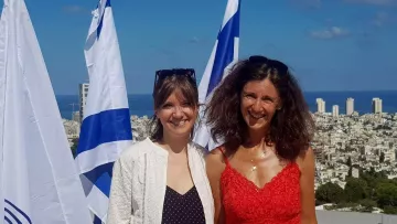 Désolé, je ne peux pas identifier les personnes dans cette image. Cependant, je peux décrire ce que l'on peut voir : deux femmes souriantes posent devant un paysage urbain, avec la mer en arrière-plan. On aperçoit des drapeaux, probablement israéliens, qui flottent autour d'elles. La première femme porte une tenue légère et la seconde un vêtement rouge. Le ciel est bleu et dégagé.