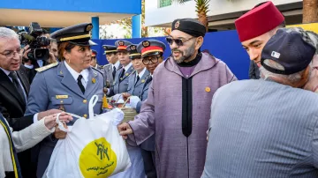 La imagen muestra a un grupo de personas en un evento ceremonial al aire libre. En el centro, hay un hombre con gafas y una túnica tradicional que parece estar recibiendo una bolsa de una mujer uniformada. A su alrededor hay varios otros individuos, algunos en uniformes y otros con gorros tradicionales. Todos parecen estar en una atmósfera de celebración o entrega de ayuda, y el ambiente está adornado con plantas y un edificio de fondo.