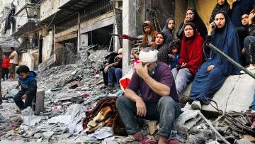 L'immagine mostra una scena devastante in un contesto di guerra o di crisi. Al centro, un uomo con una benda in testa si siede su un cumulo di macerie, con un'espressione di sofferenza e preoccupazione. Attorno a lui, un gruppo di donne sono sedute, vestite con abiti scuri e coprenti, che osservano la scena con sguardi tristi e rassegnati. Un bambino è visibile sulla sinistra, anche lui seduto tra i detriti. Il paesaggio è caratterizzato da edifici distrutti e spazzatura sparsa, evidenziando la desolazione e la disperazione della situazione.