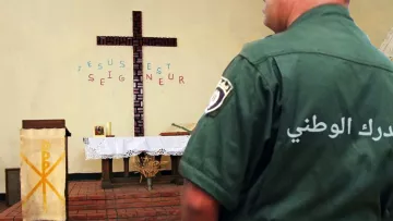 L'image montre un intérieur d'une église avec une grande croix en bois sur le mur. Des mots sont écrits autour de la croix, où l'on peut lire "JÉSUS EST SEIGNEUR". Sur la gauche, il y a une table d'autel avec un drapé. À droite, un homme en uniforme vert, probablement un agent de police ou de sécurité, est vu de dos, portant un badge qui indique "الدرك الوطني" en arabe, ce qui signifie "Gendarmerie nationale". L'ambiance de l'image semble calme et spirituelle, mêlant des éléments religieux et de sécurité.