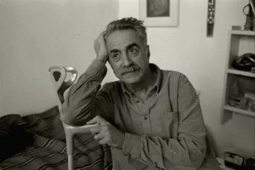 Homme pensif, assis, avec une canne, dans une pièce simple et lumineuse.