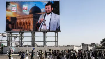 L'image montre un grand panneau d'affichage avec un portrait d'un homme en tenue traditionnelle, probablement un leader ou une figure politique, devant un paysage architectural. En dessous, un groupe de personnes marche en procession, portant des drapeaux et des affiches. L'environnement semble urbain, suggérant une manifestation ou un événement public. Les écriteaux sur le panneau en arabe ajoutent au contexte culturel et politique de l'image.