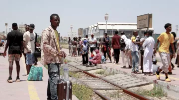 L'image montre une station de train animée. Un homme debout, tenant une valise, semble attendre. En arrière-plan, on peut voir d'autres personnes : certaines attendent assises, d'autres se déplacent avec des sacs. L'environnement est ensoleillé, et il y a des bâtiments visibles au loin. La scène évoque un moment de déplacement et d'attente.