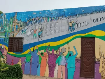 L'image montre une fresque murale colorée qui illustre une scène dynamique. On y voit des personnes célébrant et manifestant, les bras levés, sur un fond représentant un avion et une ville. Les couleurs vives ajoutent une énergie palpable à la scène, reflétant un sentiment de joie ou de solidarité. Des silhouettes de différents personnages sont visibles, et l'ensemble de l'œuvre évoque un message de rassemblement et d'espoir.