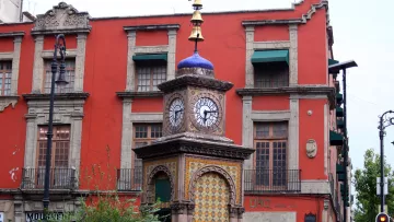 L'image montre un bel édifice, probablement une horloge monumentale, se tenant au milieu d'un espace urbain. L'horloge a une structure décorative, avec un dôme bleu et des détails en mosaïque. En arrière-plan, on peut voir un bâtiment aux murs rouges, avec des fenêtres à volets verts et des ornements architecturaux. La scène évoque une ambiance historique et charmante, typique de certaines villes anciennes. Quelques arbres et de la verdure entourent l'horloge, ajoutant une touche de nature à cet environnement urbain.