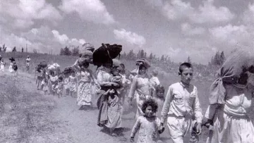 L'image montre un groupe de personnes marchant sur un chemin. On y voit des hommes, des femmes et des enfants, portant des charges sur leur tête ou dans leurs bras. Les adultes semblent laborieux, tandis que les enfants marchent à leurs côtés. Le paysage est rural, avec un ciel nuageux en arrière-plan, et l'atmosphère générale évoque une situation de déplacement ou d'exode. Les vêtements des personnes sont simples et évoquent une époque passée.