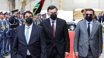 L'image montre un groupe de trois hommes marchant sur un tapis rouge, apparemment lors d'un événement officiel. Ils portent tous des costumes, et deux d'entre eux portent des masques faciaux noirs. En arrière-plan, on peut voir une garde d'honneur en uniforme, ainsi que des drapeaux. L'atmosphère semble solennelle et accueillante, suggérant une réception ou une cérémonie diplomatique.