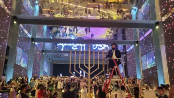 L'immagine mostra un evento festivo e affollato in un grande spazio, con una decorazione vibrante e luci colorate. Al centro, c'è una grande menorah, simbolo della tradizione ebraica, e una persona in cima a una scala che sembra essere coinvolta nella cerimonia di accensione. Il pubblico è composto da diverse persone in abiti tradizionali e moderni, tutte rivolte verso il palco. Il soffitto è riflettente, amplificando l'atmosfera festiva, mentre sullo sfondo si possono notare schermi luminosi e decorazioni artistiche. L'intera scena trasmette un senso di celebrazione e comunità.