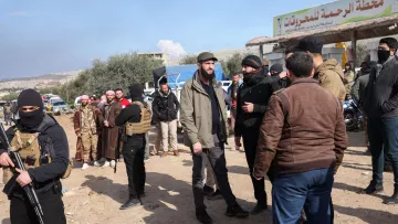 L'image montre un groupe de personnes réunies dans un environnement en plein air, vraisemblablement dans une zone rurale ou semi-urbaine. Certains hommes portent des vêtements traditionnels, tandis que d'autres sont habillés de manière plus moderne. Il y a également des individus en tenue de camouflage avec des gilets tactiques, qui semblent être armés. En arrière-plan, on peut apercevoir un panneau signalétique. Le ciel est partiellement nuageux, ce qui suggère une atmosphère de journée. Les gens interagissent entre eux, ce qui laisse penser qu'il s'agit d'une rencontre ou d'une discussion.