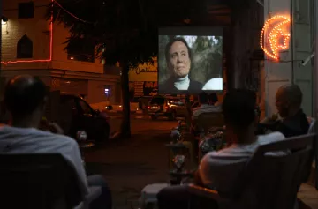 The image depicts a night scene where a group of people is seated in chairs, watching a film projected onto a large screen set up outside. The atmosphere appears relaxed, with some of the viewers enjoying drinks, possibly shisha. The area is illuminated by both the projection light and neon signage in the background, adding a lively ambiance to the setting. The scene captures a communal experience centered around outdoor movie viewing.