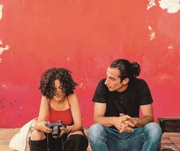 L'image montre deux personnes assises devant un mur rouge écaillé. La première personne, une femme aux cheveux bouclés, tient un appareil photo et semble concentrée sur son écran. Elle porte un débardeur rouge et un gilet clair. À côté d'elle, un homme avec des cheveux longs attachés en chignon la regarde attentivement. Il est vêtu d'un t-shirt noir et de jeans. L'atmosphère paraît détendue et intime, comme s'ils étaient en pleine conversation ou en train de partager un moment.