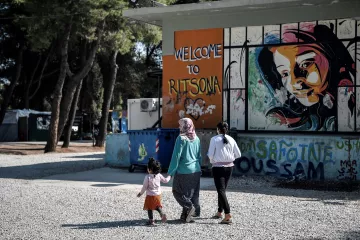 L'immagine mostra tre donne che stanno camminando in una zona all'aperto. Una di loro tiene per mano una bambina, mentre un'altra donna indossa un hijab. Sullo sfondo si può vedere un murale colorato che rappresenta un volto di donna, accompagnato dalla scritta "Welcome to Ritsona". L'area sembra essere informale e accogliente, con alberi che la circondano e strutture varie visibili.