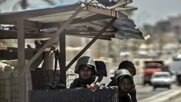 The image depicts a group of soldiers stationed at a military outpost. They are wearing helmets and carrying weapons, suggesting they are on alert or guarding a specific area. The backdrop includes palm trees and some vehicles, indicating an urban environment. A flag can be seen on top of the structure, adding to the military context of the scene. The overall atmosphere appears tense and focused on security.