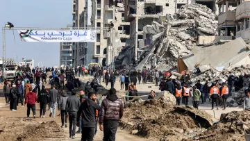 L'image montre une scène dans une ville en reconstruction, probablement à Gaza. On voit des bâtiments partiellement détruits, avec des débris éparpillés. De nombreuses personnes marchent dans la rue, certaines portant des gilets de bénévoles ou de secours. Un panneau en arabe et en anglais indique "Bienvenue à Gaza". L'ambiance dégage un mélange d'espoir et de désolation, avec des gens qui semblent se diriger vers un lieu particulier. Les conditions environnantes sont poussiéreuses et témoignent des récents conflits.